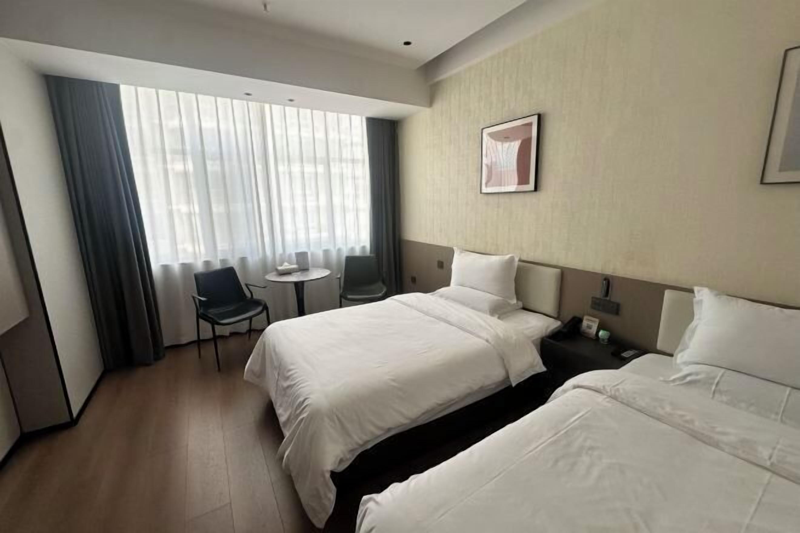 A hotel room with two beds, a small table, a chair, and a window with curtains.