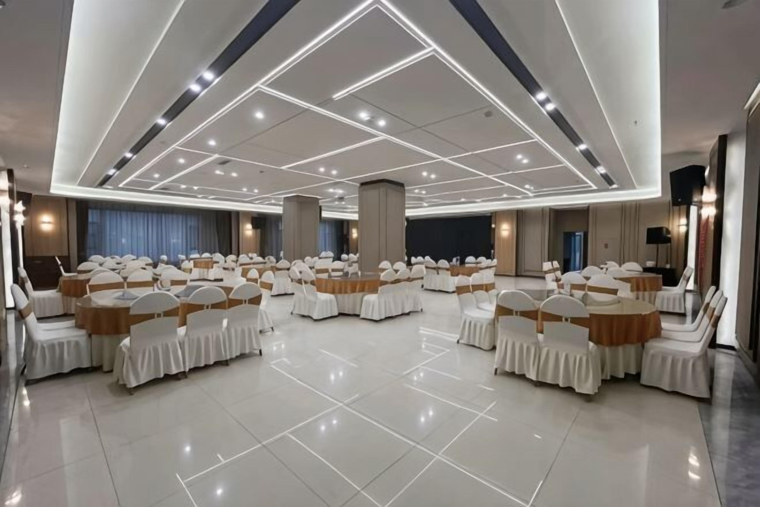 Une grande salle de conférence, bien éclairée, avec des rangées de tables et de chaises, un plafond haut doté d’un éclairage géométrique et un espace dédié à la scène.