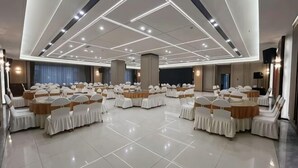Une grande salle de conférence, bien éclairée, avec des rangées de tables et de chaises, un plafond haut doté d’un éclairage géométrique et un espace dédié à la scène.