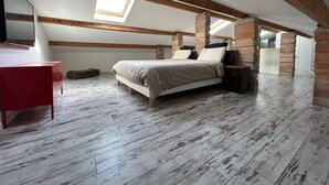 Ein Schlafzimmer mit geneigter Decke, freiliegenden Holzbalken, einem Oberlicht, einem Bett mit Kissen, einem Nachttisch und einem roten Schreibtisch.