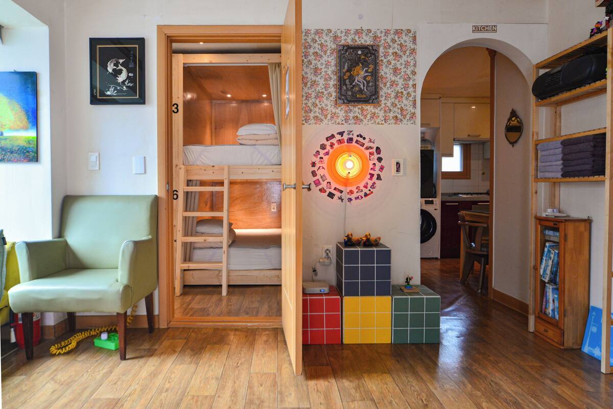 Ein Zimmer mit einem Etagenbett, einem grünen Sessel, einem bunten Spielbereich und einer durch einen Bogen sichtbaren Küche.