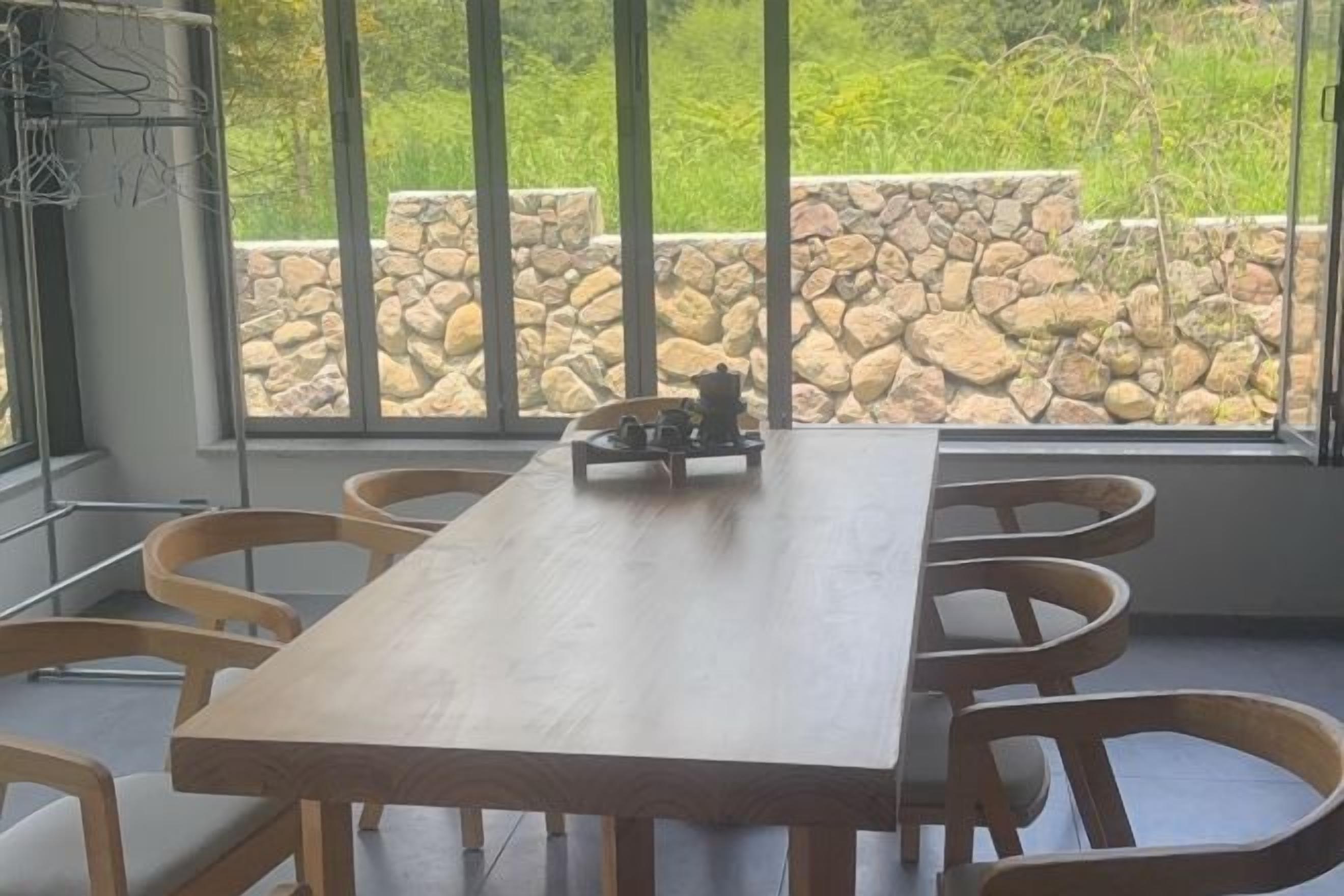 A wooden dining table with six chairs, large windows with a stone wall view, and a metal rack with clothes.