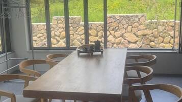 A wooden dining table with six chairs, large windows with a stone wall view, and a metal rack with clothes.