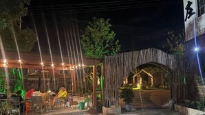 An outdoor seating area with tables and chairs, a covered section with string lights, and a wooden archway leading to a lit pathway. - Anji Qile Farm (Anji)