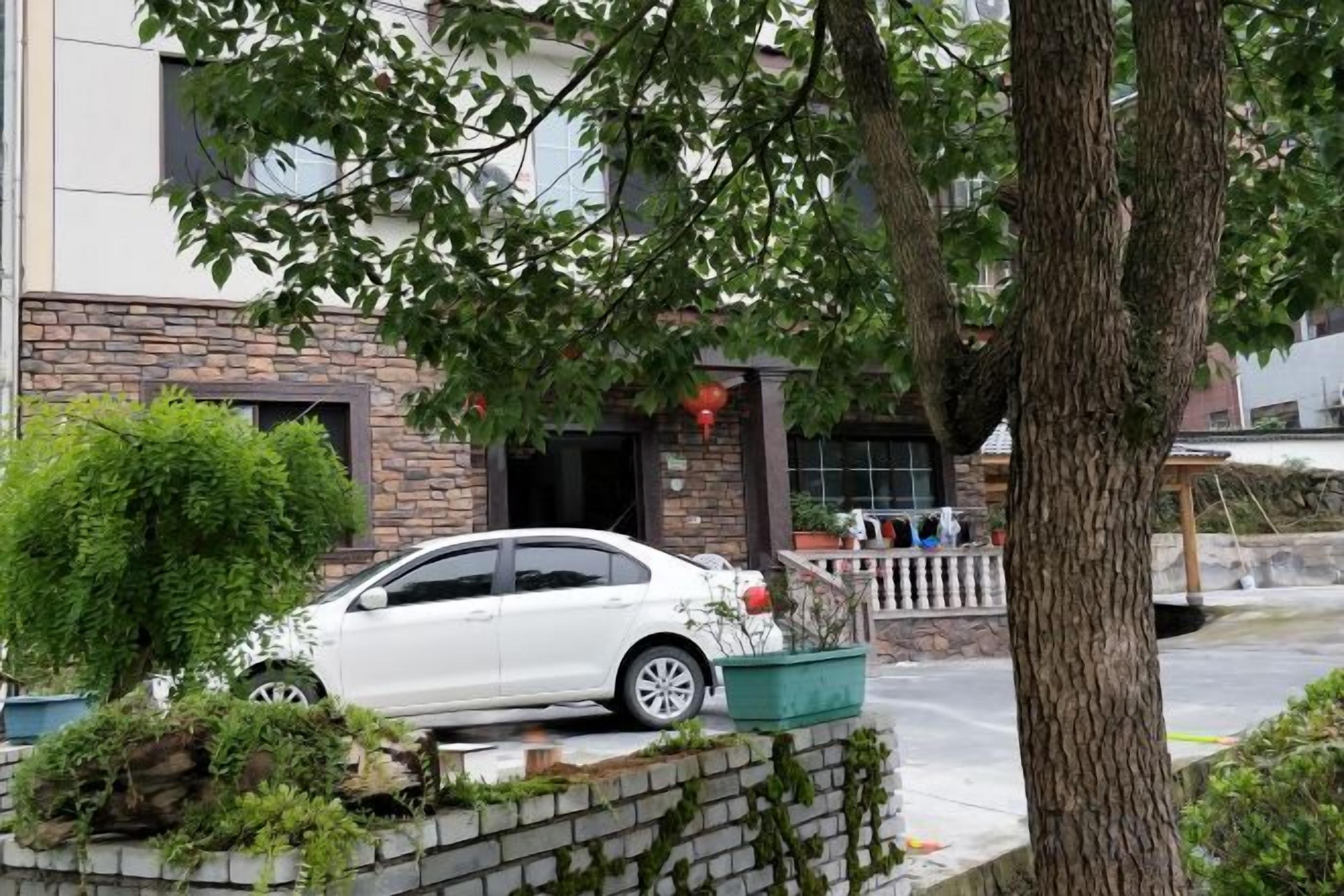 Um carro branco estacionado em frente a um edifício com fachada de tijolos, uma árvore e uma pequena área ajardinada.