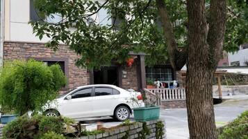 Um carro branco estacionado em frente a um edifício com fachada de tijolos, uma árvore e uma pequena área ajardinada.