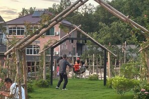 Een tuin met een houten constructie, mogelijk een pergola, omgeven door groen en een huis op de achtergrond.