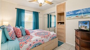 A bedroom with a bed, a ceiling fan, a TV, and a painting of a beach scene. - Long Island Village 314 (Port Isabel)