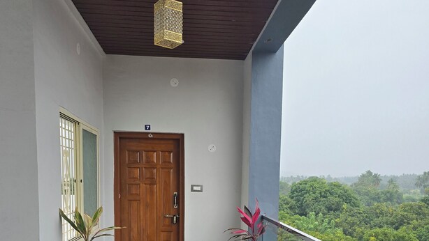 A balcony with a view of greenery, a wooden door, and potted plants. - Lakshmi Villa (Hospet)