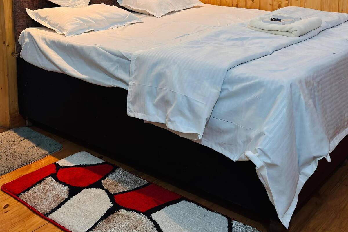 Un dormitorio con cabecera de madera, cama con ropa de cama blanca, alfombra estampada en rojo y blanco, y dos luces fijadas a la pared.
