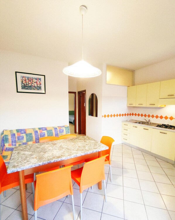 A dining area with a table and chairs, a hanging light fixture, and a kitchen with yellow cabinets. - Bright and Comfortable Flat (San Michele al Tagliamento)