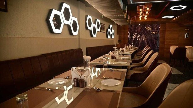 A restaurant with long tables, brown leather booths, and hexagonal wall decorations. - Hotel Baba Vishwanath Palace  (Prayagraj)