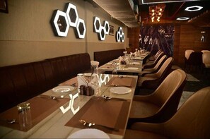 A restaurant with long tables, brown leather booths, and hexagonal wall decorations. - Hotel Baba Vishwanath Palace  (Prayagraj)