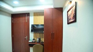 A compact room with a wooden wardrobe, a small desk with a chair, a microwave, and a framed picture on the wall. - Hotel Baba Vishwanath Palace  (Prayagraj)