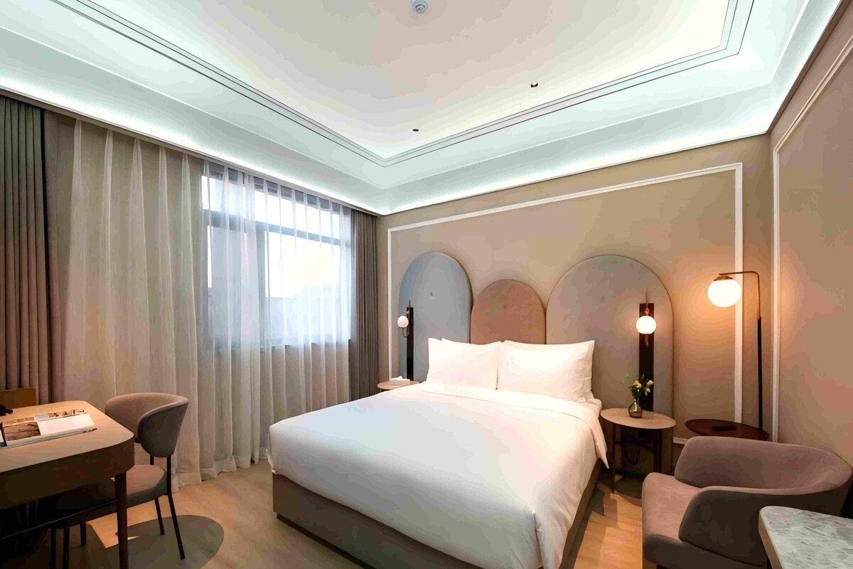A modern hotel room with a large bed, a desk, two chairs, and a nightstand with a lamp.