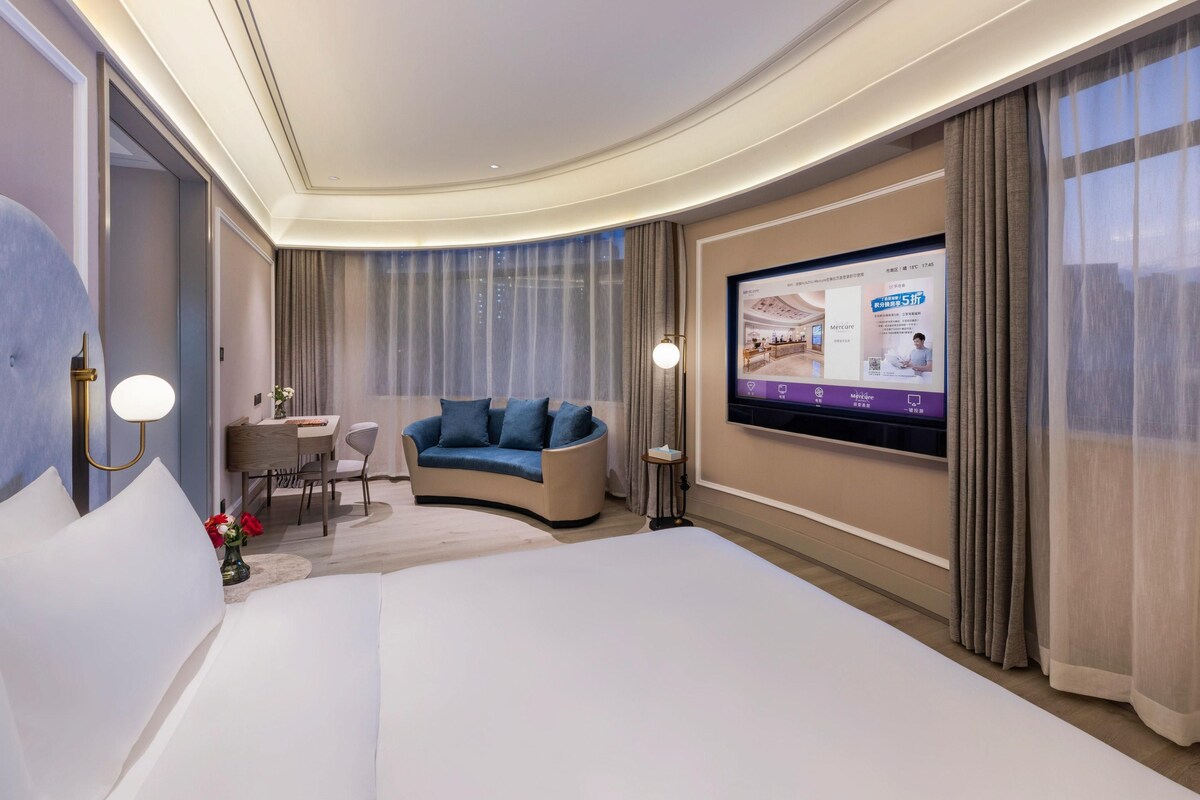 A hotel room with a large bed, a sofa, a desk with a chair, a TV mounted on the wall, and a large window with curtains.