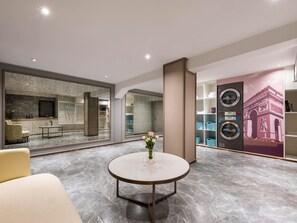 Un moderno vestíbulo de hotel con suelo de mármol, una mesa redonda con un jarrón, una zona de estar, un espejo grande y una pared con un mural decorativo.