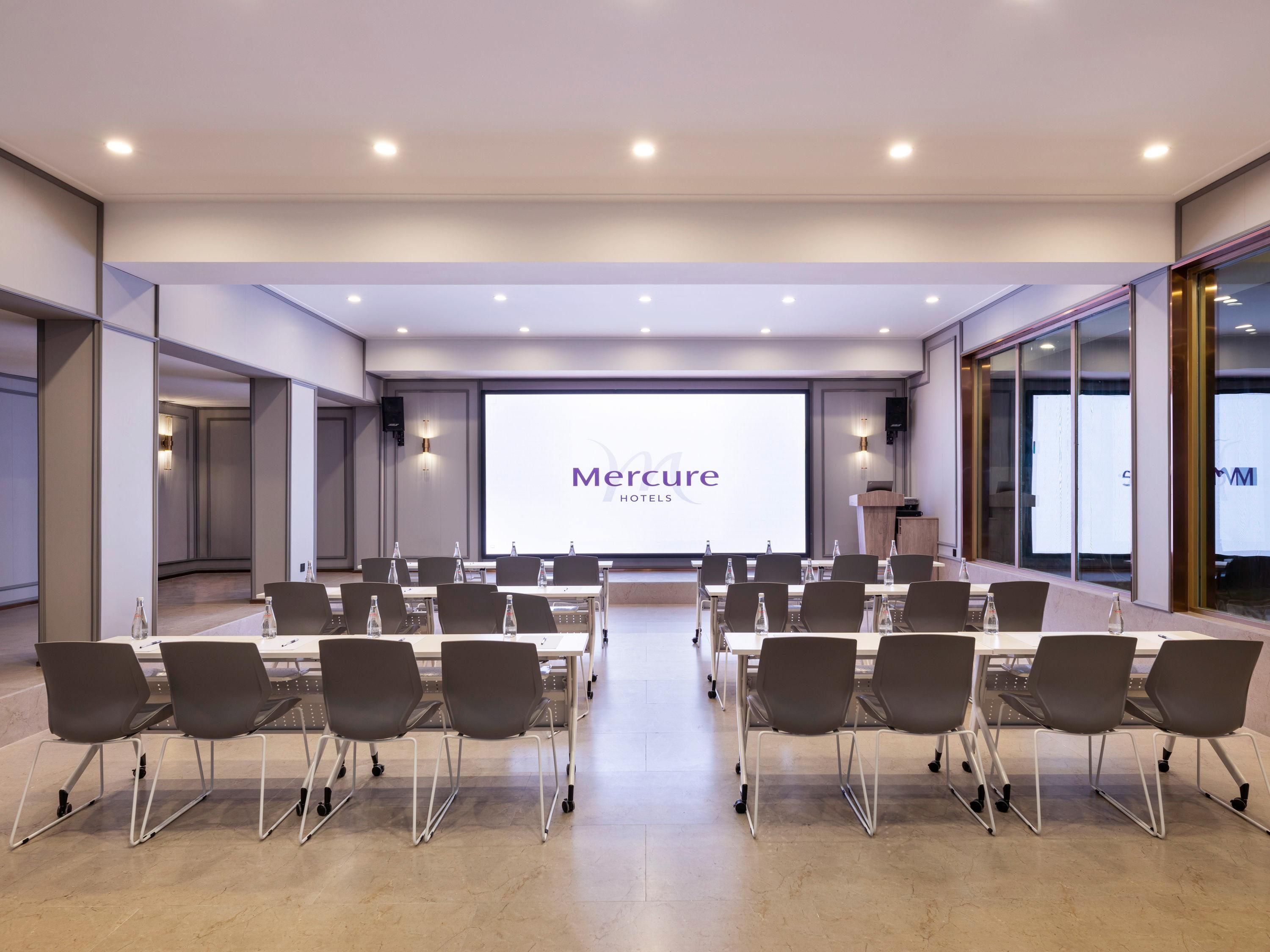 A conference room with a large screen displaying 'Mercure Hotels' branding, equipped with multiple tables and chairs.