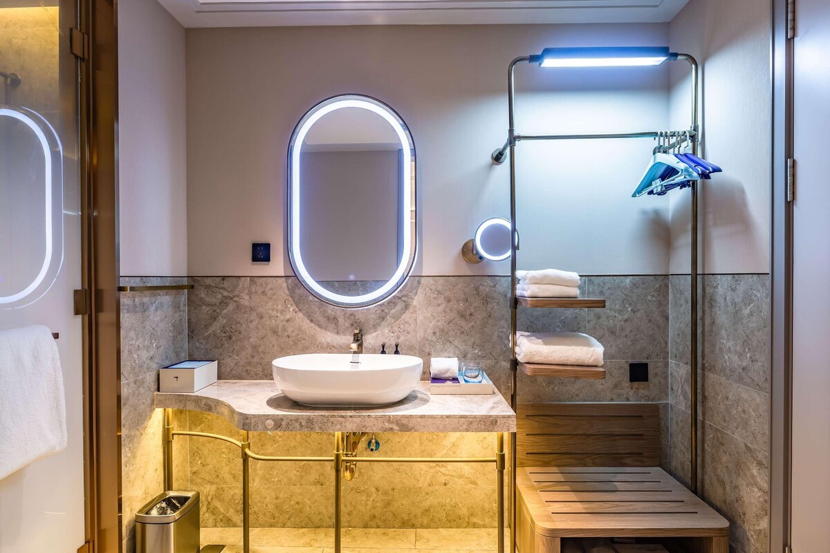 A modern bathroom with a vanity, a large mirror, a shower area, and a wooden bench.