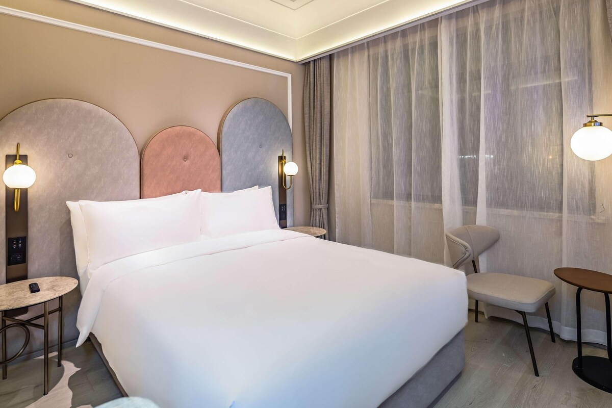 A hotel room with a large bed, a small round table, a chair, and a desk.