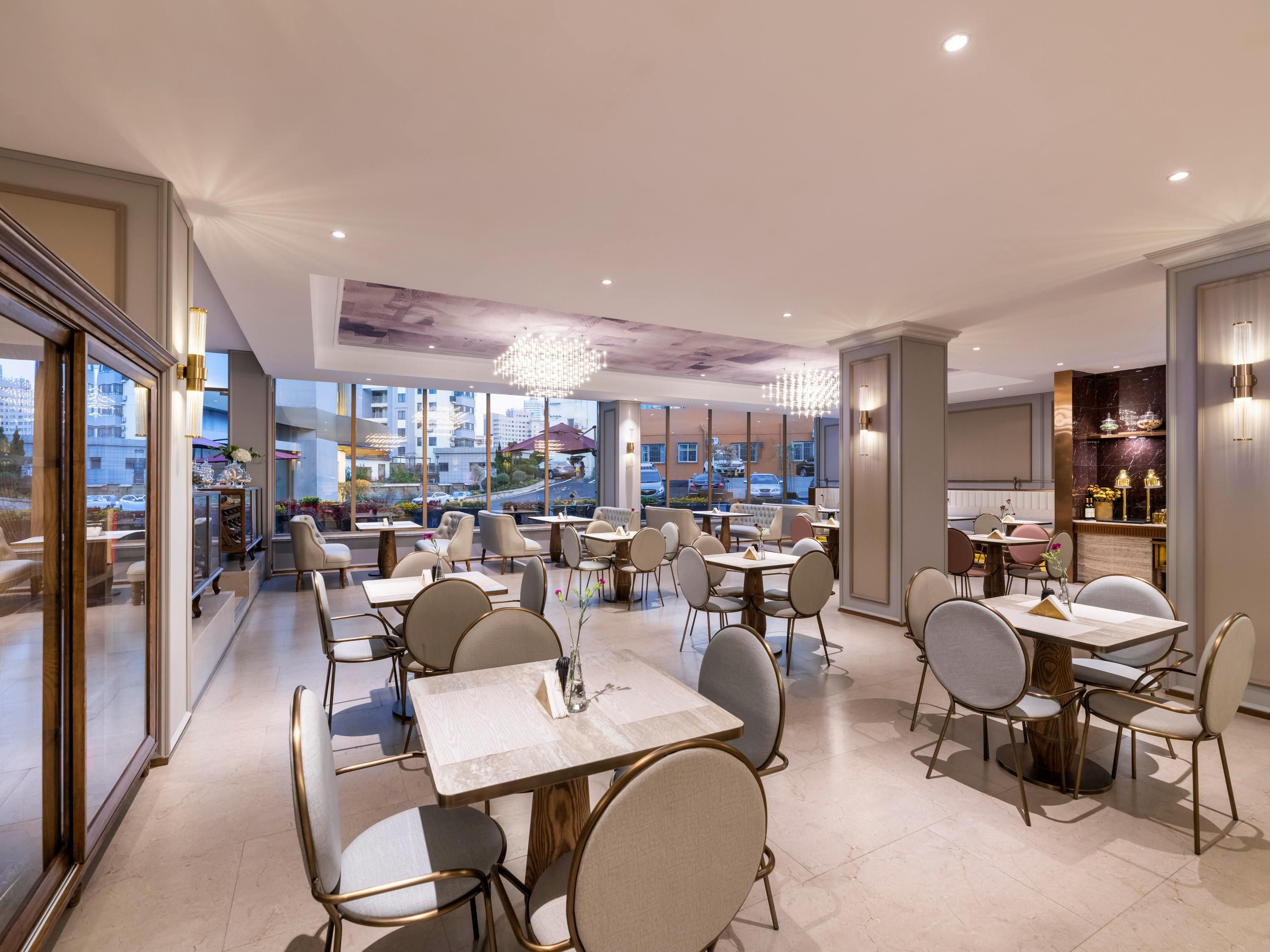 A modern restaurant with multiple dining tables and chairs, large windows offering city views, and elegant lighting fixtures.
