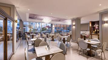 A modern restaurant with multiple dining tables and chairs, large windows offering city views, and elegant lighting fixtures.