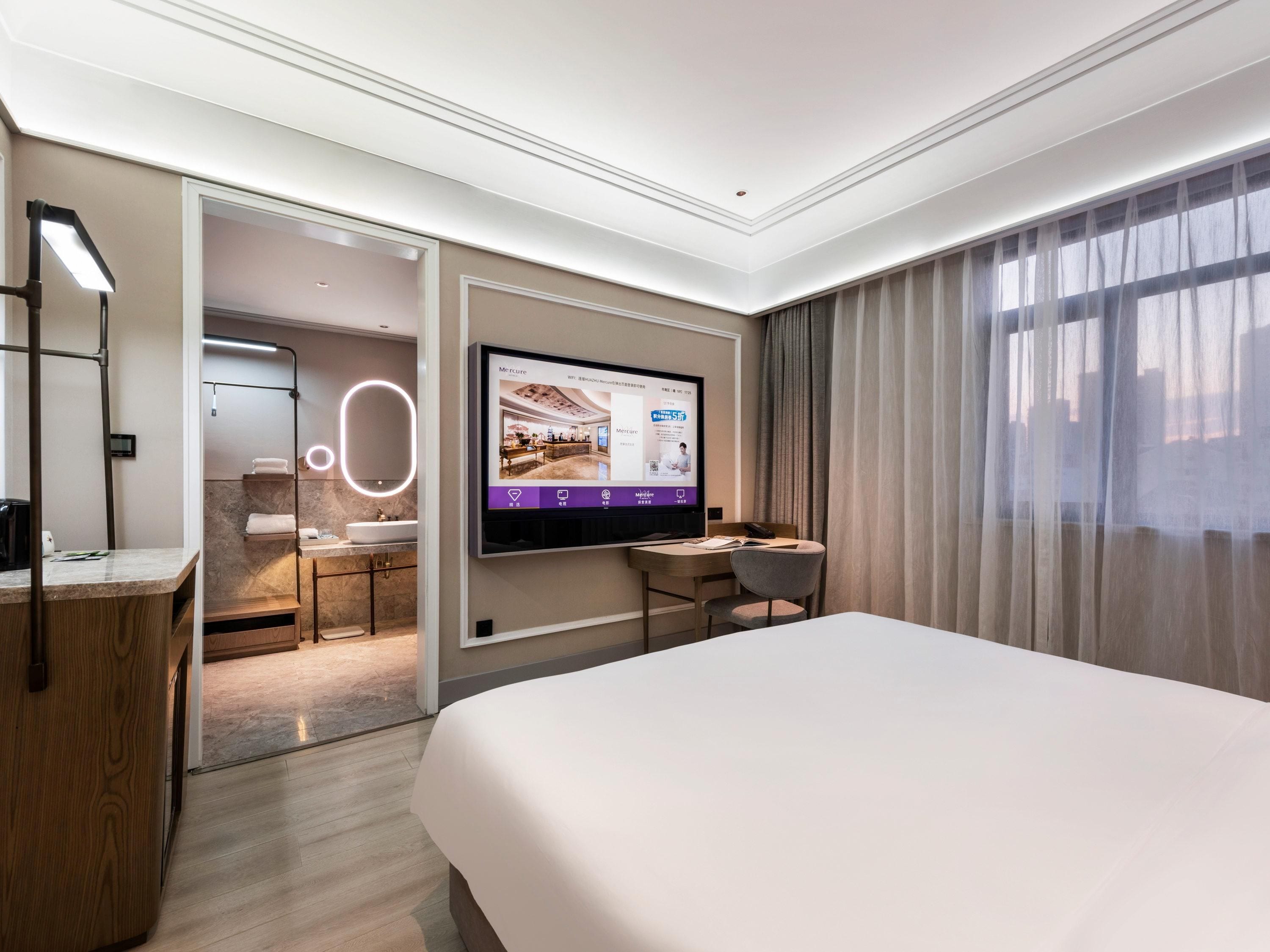 A modern hotel room with a large bed, a desk, a TV mounted on the wall, and a bathroom visible through an open door.