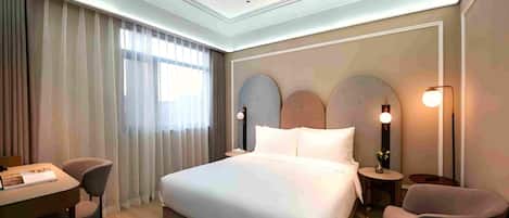 A modern hotel room with a large bed, a desk, two chairs, and a nightstand with a lamp.