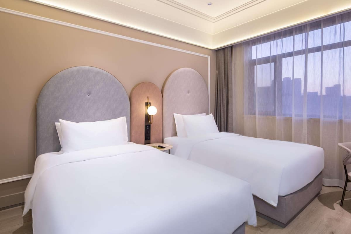 Una habitación de hotel con dos camas, una mesita de noche con lámpara, una ventana con cortinas y vistas a la ciudad.