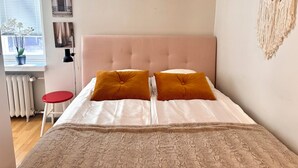 A bedroom with a pink tufted headboard, two mustard pillows, a white bedspread, a red stool, a window with blinds, a radiator, and a macrame wall hanging.