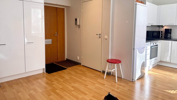 A modern kitchen with white cabinets, a wooden door, a red stool, and a built-in oven.