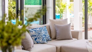 A modern living room with a beige sofa, a patterned pillow, and a view of a pool through large windows. - Bali Style Pool Villa - Private & Serene (Choeng Mon)