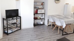Un espacio habitable compacto con un televisor sobre un soporte negro, una mesa de comedor con sillas, una zona de cocina con microondas y estantes, y un pequeño pasillo.