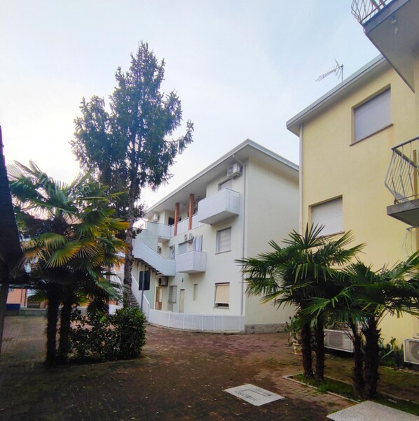 Un immeuble résidentiel à plusieurs étages, doté de balcons, d’une cour pavée et entouré de palmiers.