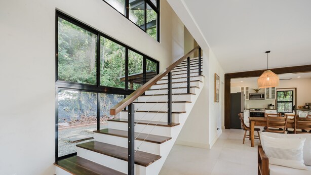 Un intérieur moderne avec un escalier menant à un deuxième étage, un coin repas avec des meubles en bois et de grandes fenêtres offrant une vue sur la verdure.
