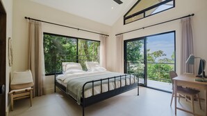 Une chambre à coucher avec un lit, un bureau et une chaise. De grandes fenêtres offrent une vue sur la verdure environnante.