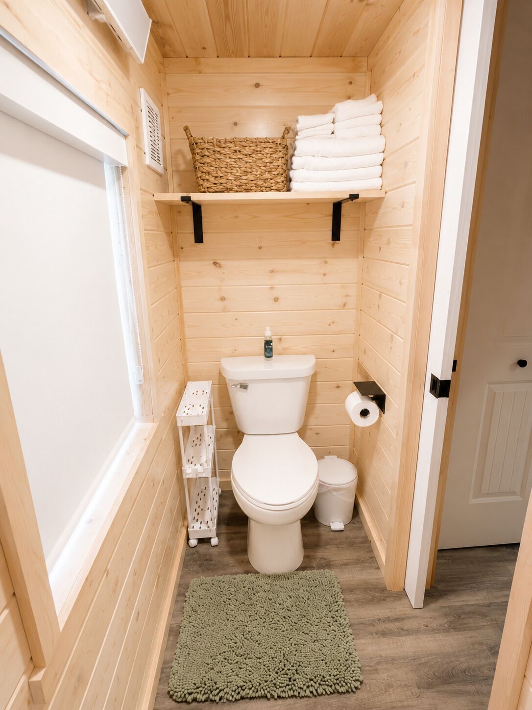 Une petite salle de bain en bois, équipée de toilettes, d’une petite étagère avec des serviettes, d’un panier et d’un tapis vert.