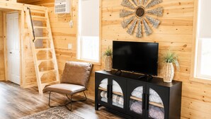 A wooden interior with a TV on a black cabinet, a brown chair, a decorative wall piece, and a ladder. - Private Tiny Cabin in Glendive Near Makoshika Park - Free Parking & WiFi (Glendive)