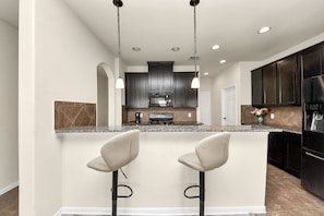A modern kitchen with dark cabinets, a central island with bar stools, and a stainless steel refrigerator. - Cozy & Spacious Getaway with King bed and Backyard (Houston)