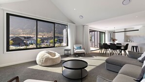 Ein modernes Wohnzimmer mit großem Fenster, einem weißen Sitzsack, einem grauen Sofa, einem Essbereich mit Tisch und Stühlen sowie einer Küche mit weißen Schränken.