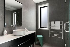 Un baño moderno con un espejo grande, un lavabo redondo, una cabina de ducha de vidrio y un taburete pequeño.