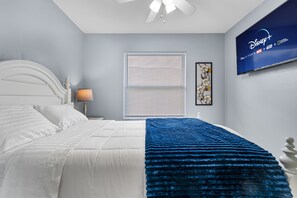 Un dormitorio con cama, televisor montado en la pared, ventana con persianas y mesita de noche con lámpara.