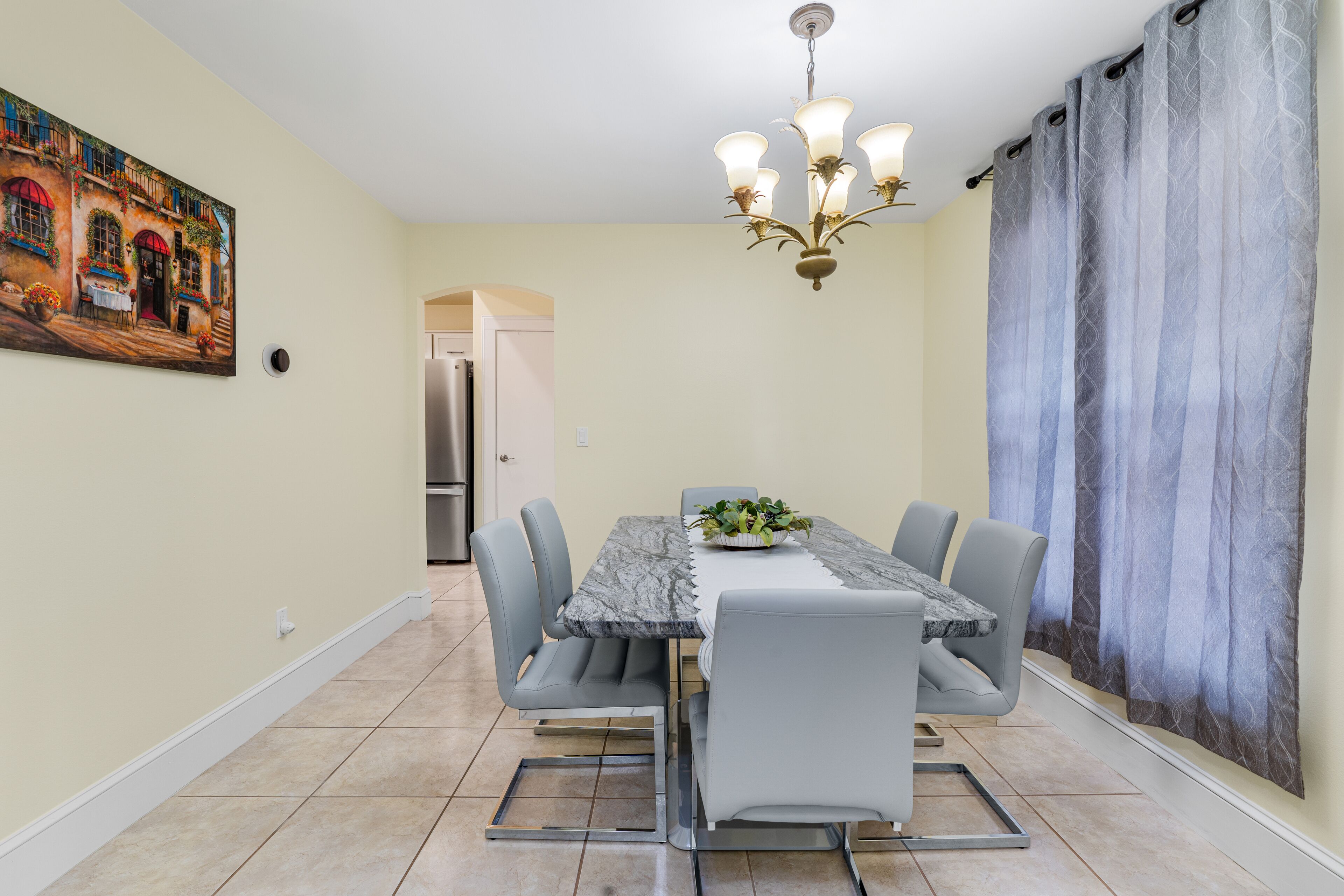 A dining area with a marble table, grey chairs, a chandelier, and a painting on the wall.