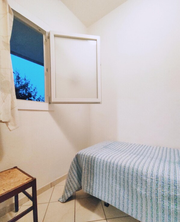 A small, single-bed room with a window, a chair, and a tiled floor.
