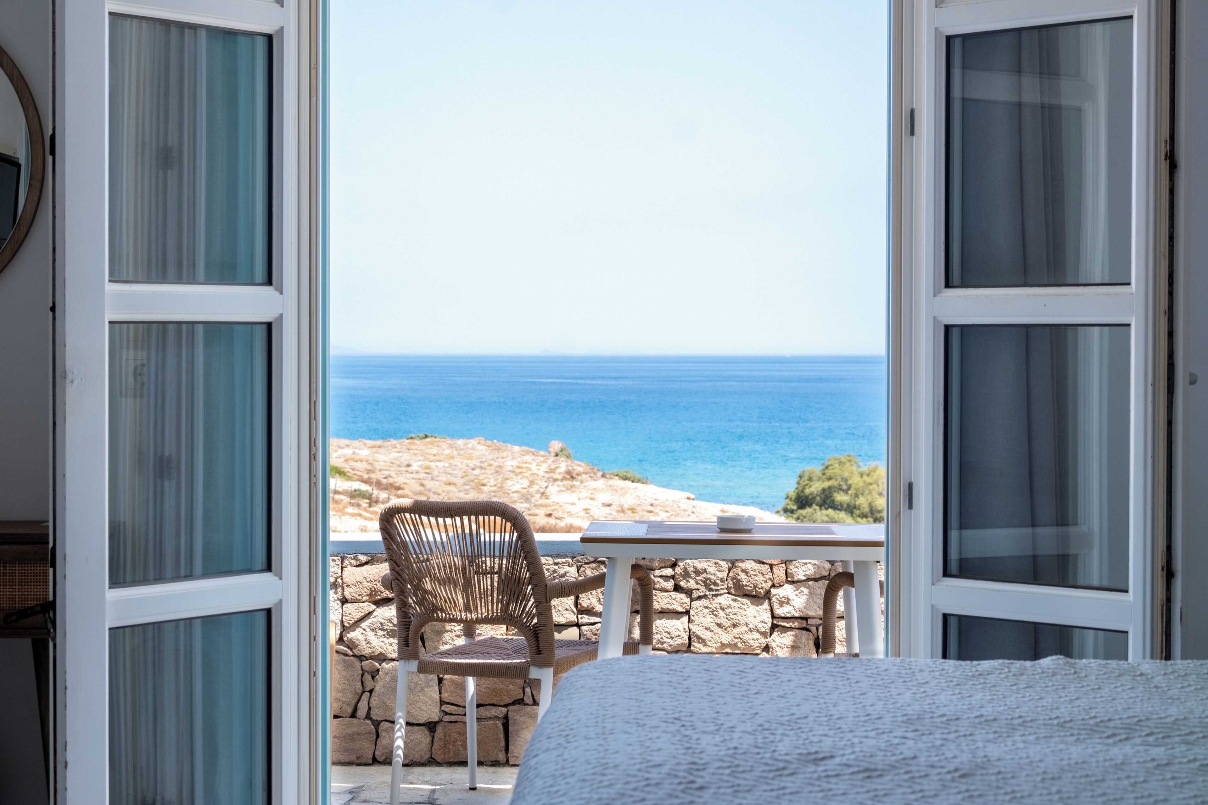 Une pièce avec un balcon ouvert, une chaise en osier, un mur de pierre et une vue sur la mer.