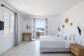 Een slaapkamer met een bed, nachtkastje en uitzicht op de zee door een open deur.