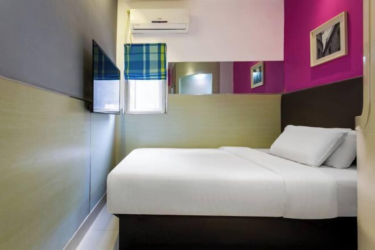 A hotel room with a bed, a window with a plaid curtain, an air conditioning unit, and a wall-mounted TV.