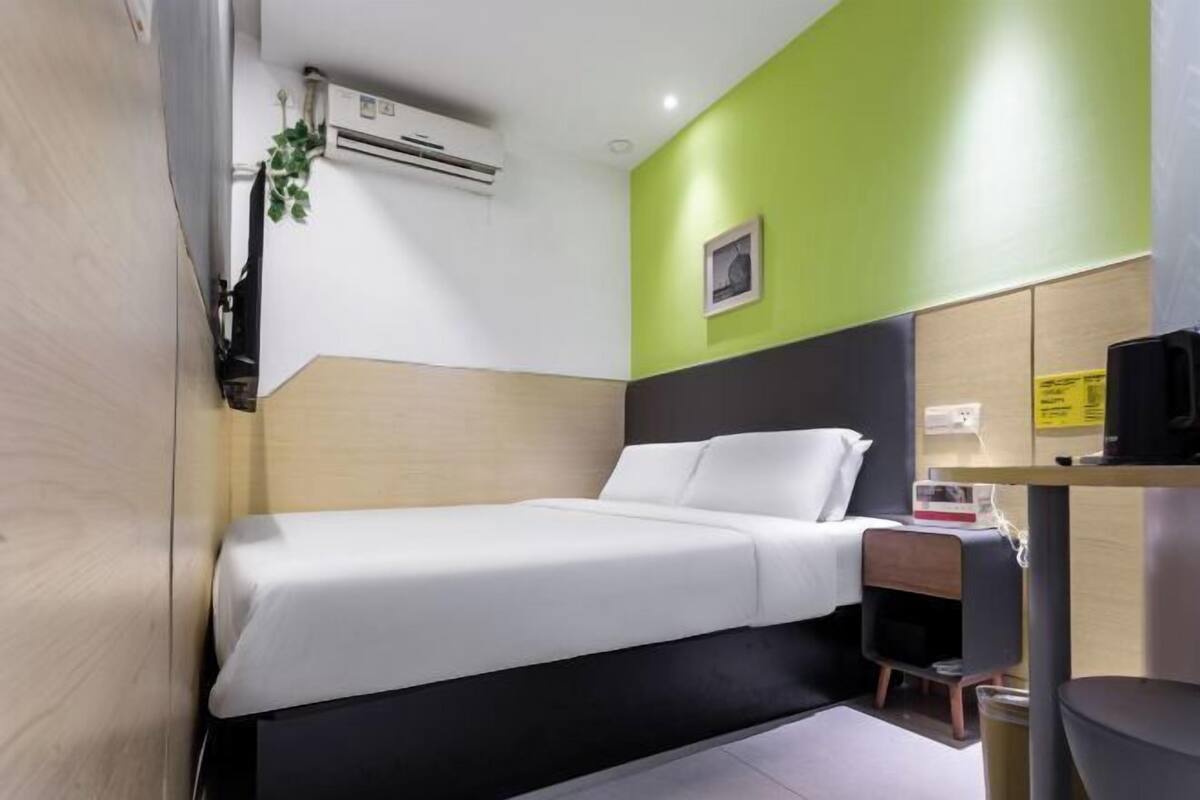 A single bed with a white mattress and pillows, a small bedside table with a lamp, a wall-mounted air conditioner, and a green accent wall.