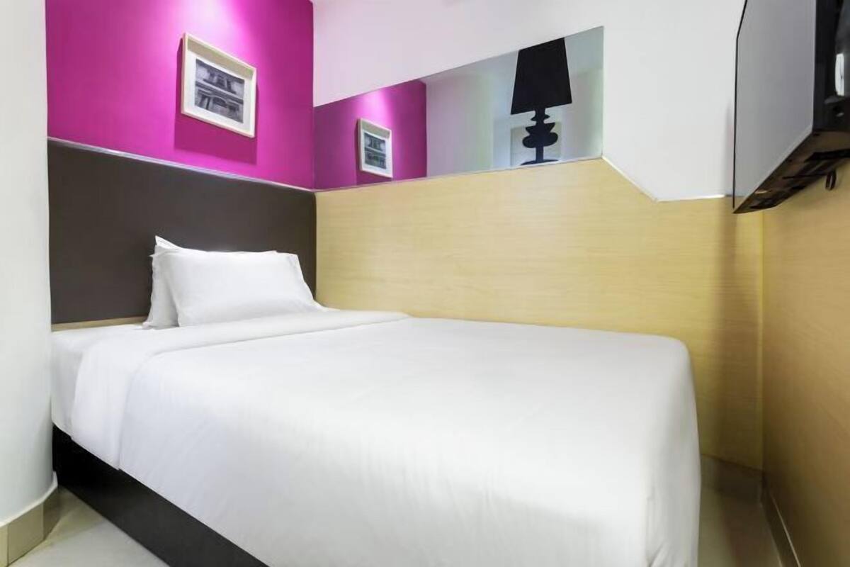 A hotel room with a bed, a headboard, a wall-mounted lamp, and framed pictures.