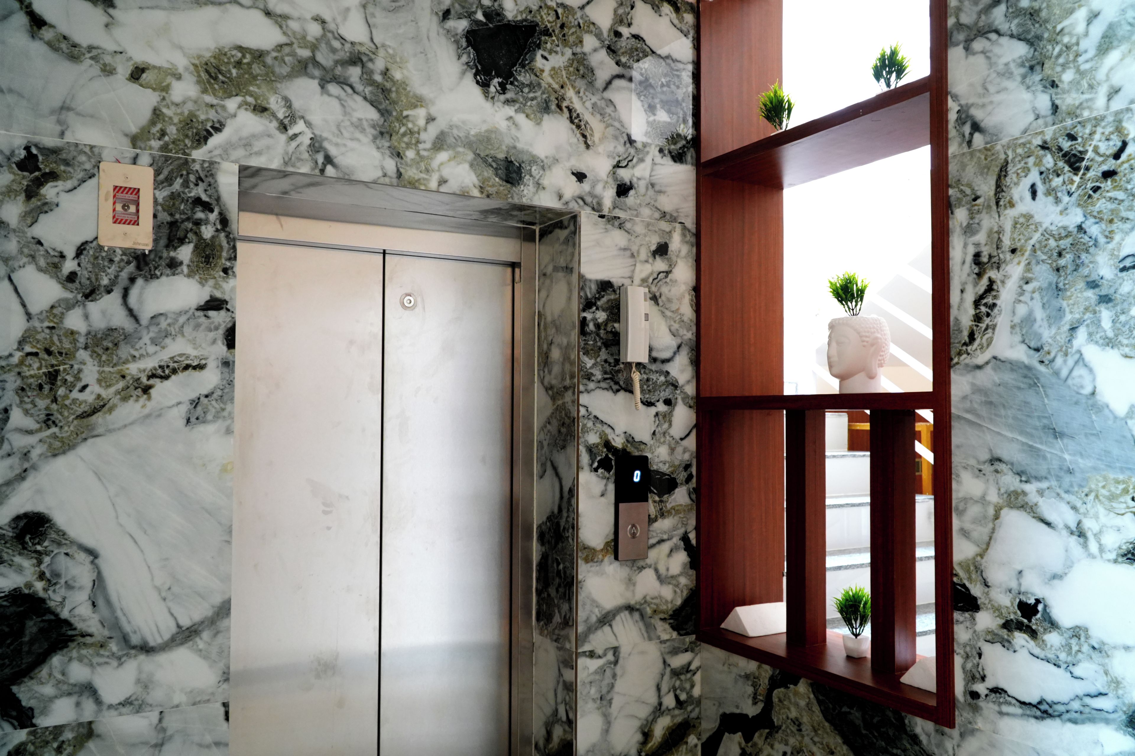An elevator with a marble wall, a wooden shelf with plants, and a small sign on the wall.